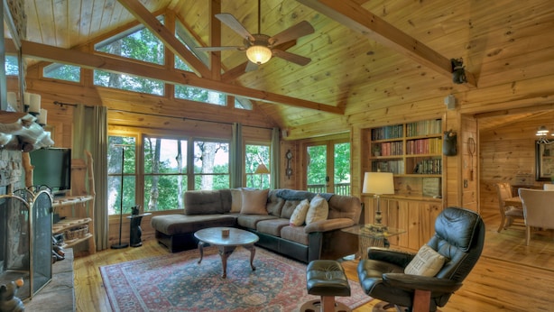 A cozy cabin interior with a wooden ceiling, large windows, a sectional sofa, a leather armchair, and a bookshelf filled with books. - Stunning Mountain Cabin w/ Pool Table, Hot Tub, Wifi, 20 min to Blue Ridge (Blue Ridge)