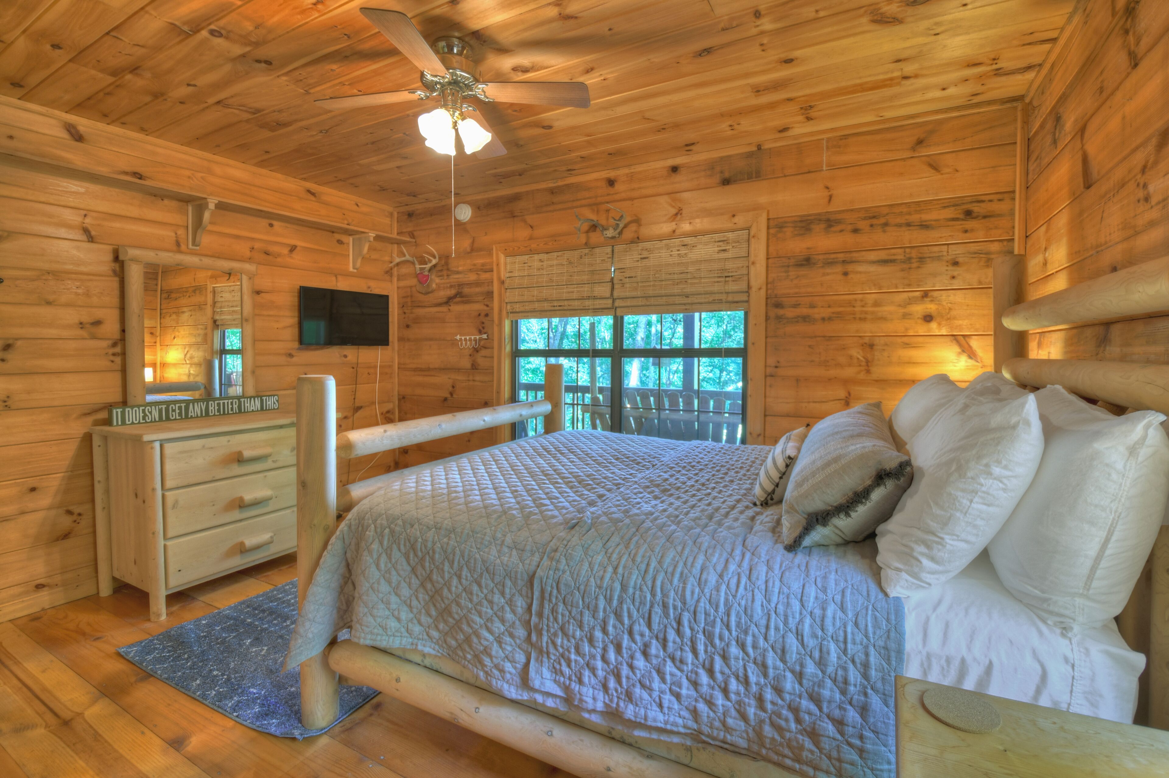 A wooden cabin bedroom with a bed, dresser, television, and a window with a view of trees.