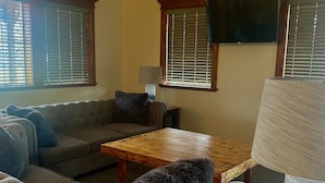 Ein Wohnzimmer mit einer Couch, einem Holztisch, einer Lampe und einem Fernseher an der Wand.