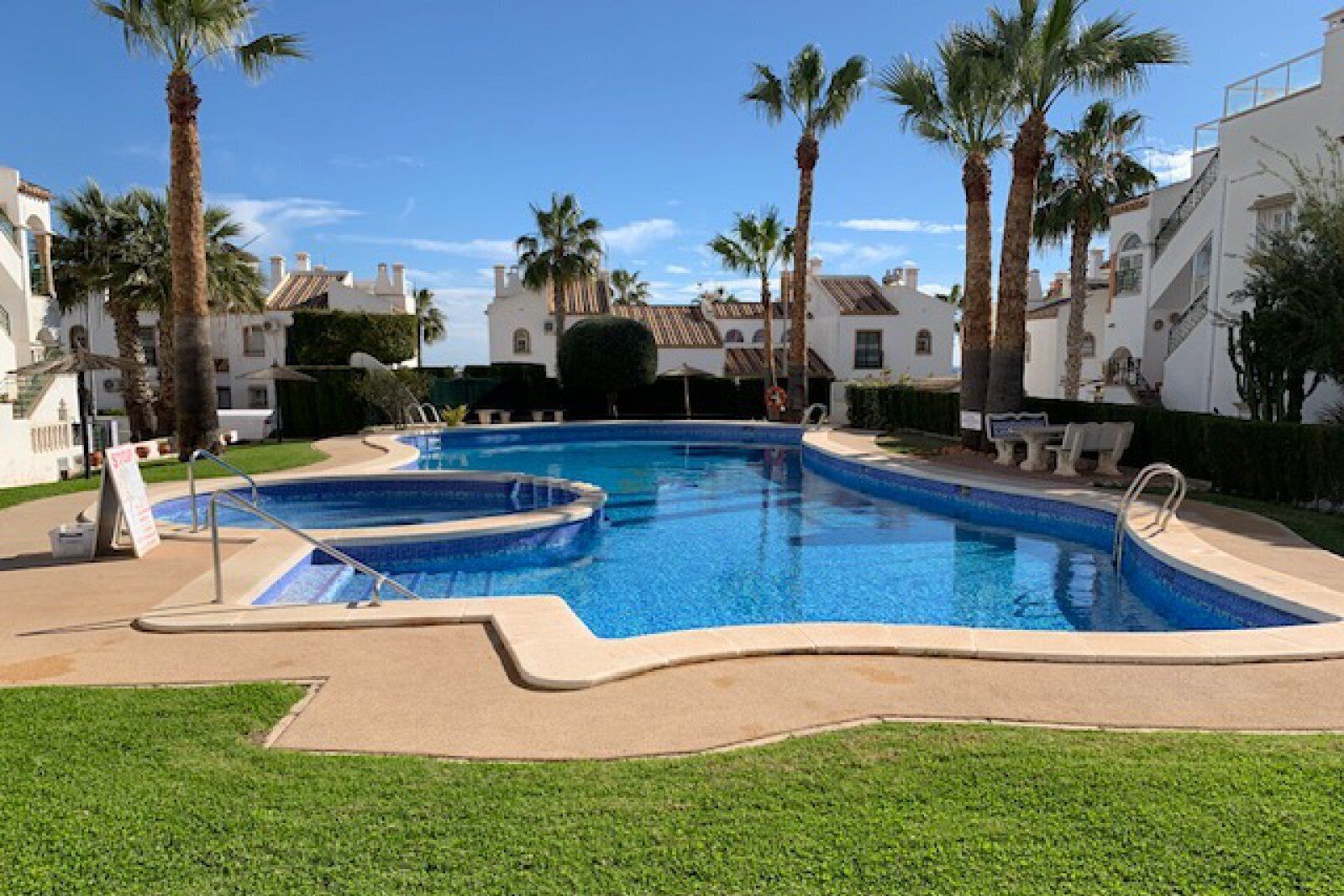 A swimming pool with a pool ladder, surrounded by palm trees and a white building with a tiled roof.