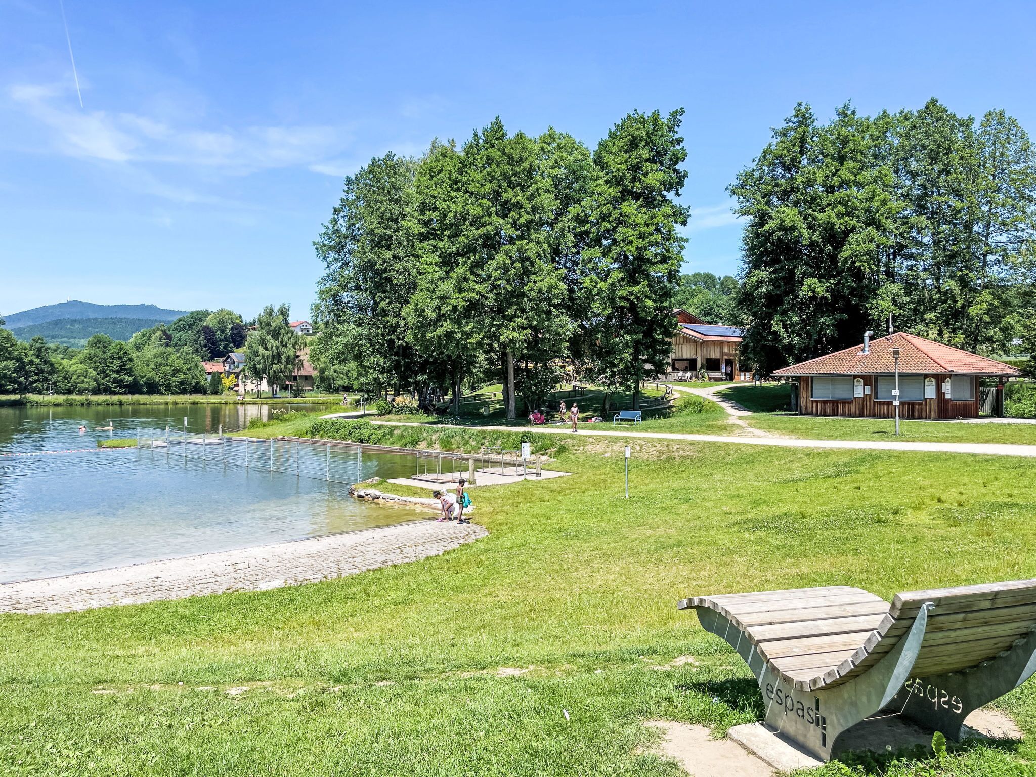 Un parc au bord du lac, avec un banc en bois, un petit bâtiment et un ciel dégagé.