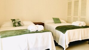 A hotel room with two beds, each with white linens and a green throw blanket. There are pillows on the beds, and a green pillow with text on it. The room has a light-colored wall and a set of white lockers in the background.