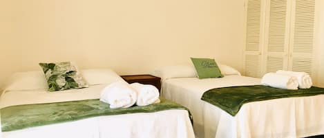 A hotel room with two beds, each with white linens and a green throw blanket. There are pillows on the beds, and a green pillow with text on it. The room has a light-colored wall and a set of white lockers in the background.