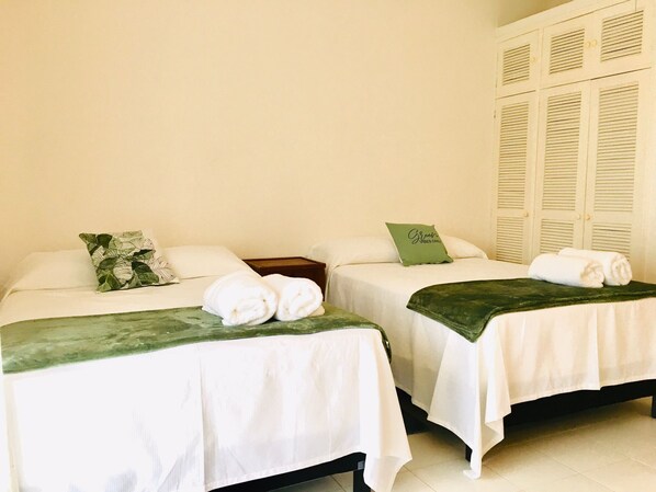 A hotel room with two beds, each with white linens and a green throw blanket. There are pillows on the beds, and a green pillow with text on it. The room has a light-colored wall and a set of white lockers in the background. - House buganvilias in hotel zone Cancún, with pool (Cancún)