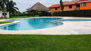 A swimming pool with a thatched-roof structure, surrounded by a well-maintained lawn and a building with a red roof.