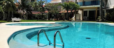 A swimming pool with a curved edge, surrounded by palm trees and a building with a red-tiled roof.