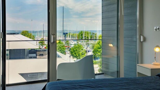 A bedroom with a large sliding glass door leading to a balcony, a bed, a chair, and a bedside table with a lamp. - Apartment Fűzfő by Interhome (Balatonszemes)