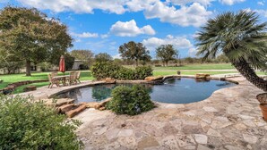 A stone pool with a surrounding patio, outdoor furniture, and a view of a golf course under a clear sky. - Historic 1906, 15 acre Ranch with pool 20 minutes from SeaWorld (San Antonio)