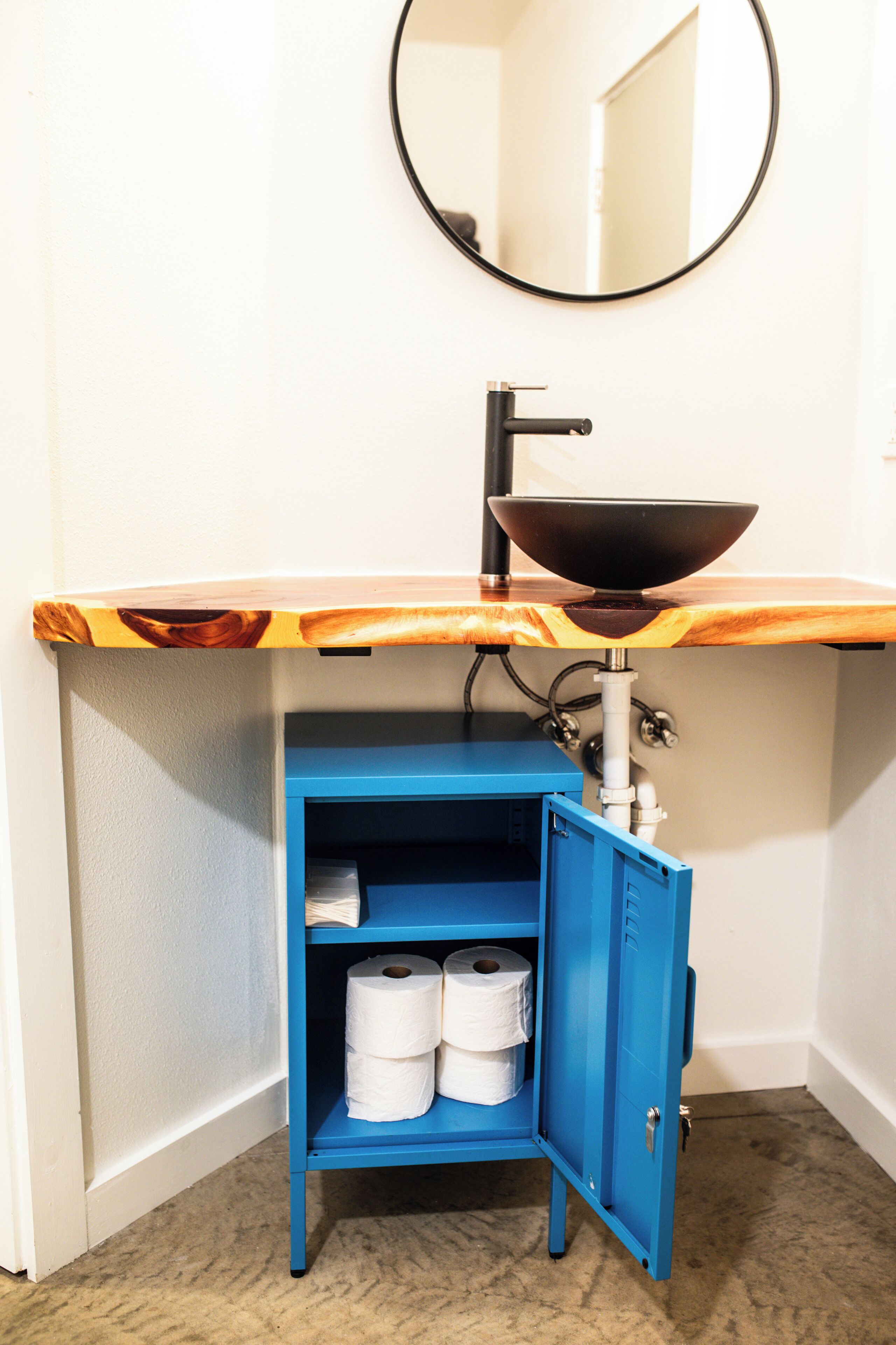 A bathroom with a wooden vanity, a black sink, and a blue cabinet with toilet paper.