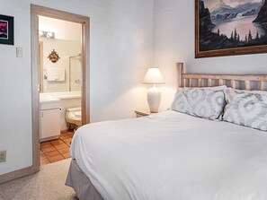 Une chambre à coucher avec un lit, une table de chevet, une lampe, un tableau, et une salle de bain visible à travers une porte ouverte.