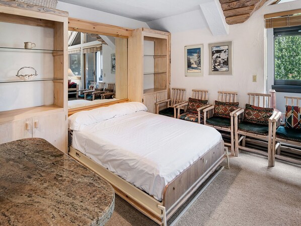 Une chambre à coucher avec un lit à baldaquin en bois, un coin salon avec des fauteuils et des coussins, et une grande fenêtre donnant sur la forêt.