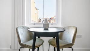 Una habitación con una mesa redonda y dos sillas, un jarrón sobre la mesa, y una ventana con vistas a edificios.