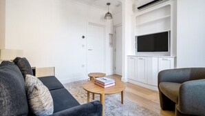Una sala de estar moderna con un sofá negro, un sillón gris, una mesa de centro de madera con libros, un televisor sobre un mueble blanco y un equipo de aire acondicionado en la pared.