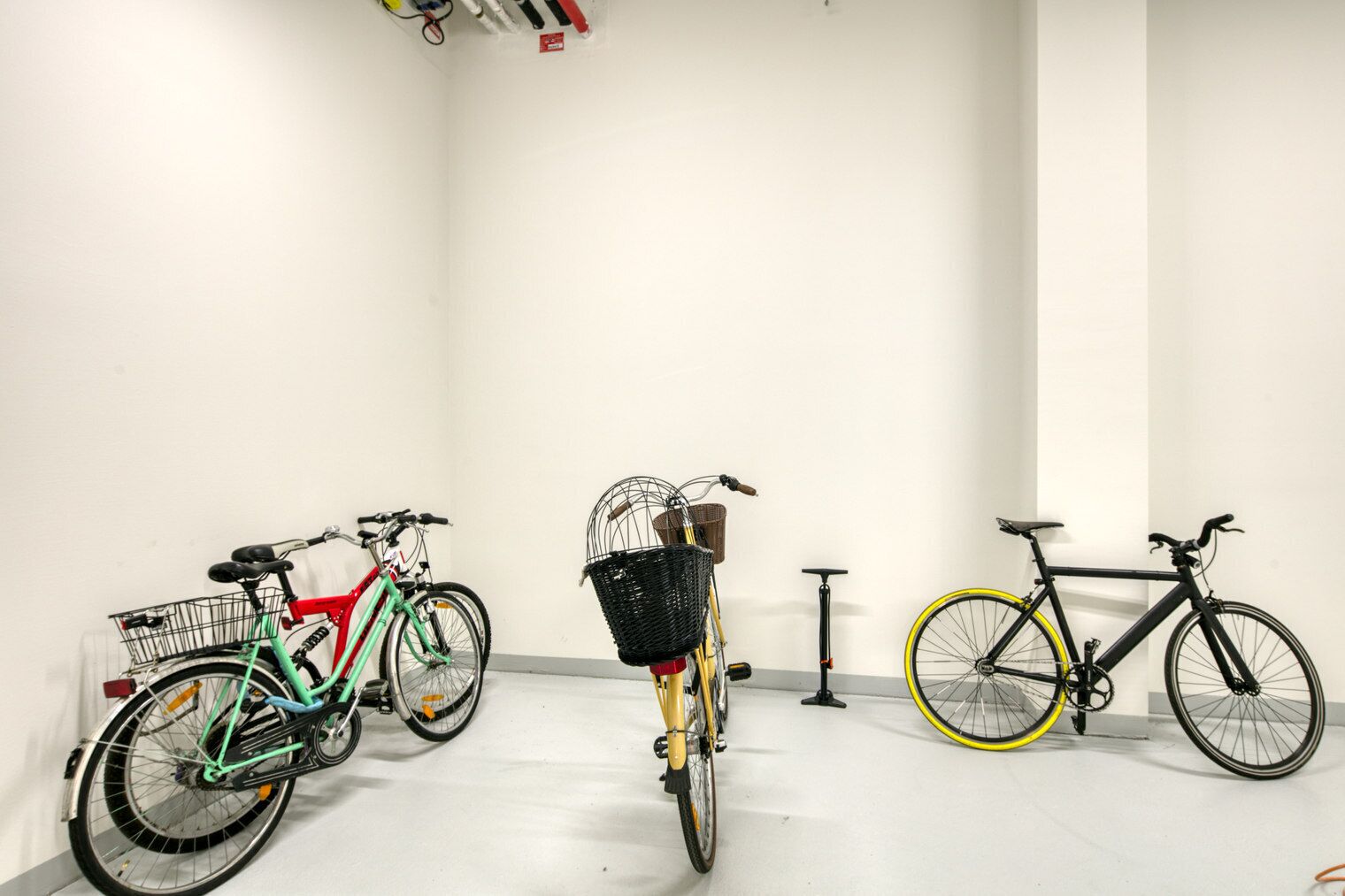 A clean, white-walled room with three bicycles parked against the wall, each with a basket or seat.
