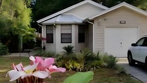 Une maison de plain-pied avec un garage, entourée d’un jardin où poussent différentes plantes, et une fleur rose au premier plan.