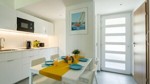 A modern kitchen with white cabinets, a black microwave, and a coffee maker. There is a dining table with blue plates and a yellow tablecloth. A painting of a sailboat is on the wall, and a door with frosted glass panels is visible. - Földvár by Interhome (Balatonszemes)