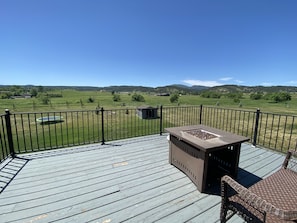 Una terraza de madera con barandilla metálica, un hogar y vistas a un campo verde y montañas.