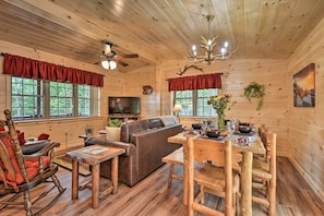 A cozy cabin interior with a wooden floor, a sofa, a dining table with chairs, a television, and a chandelier. - Secluded Mountain Cabin in Wardensville! (Wardensville)
