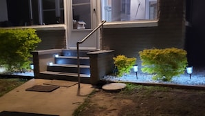 A nighttime view of a house with a front porch, steps leading to a door, and a small garden area with shrubs and a pathway. - Pensacola Breeze/ Downtown (Pensacola)