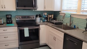 A modern kitchen with white cabinets, a stainless steel microwave, an oven, and a dishwasher. There is a window with blinds, a sink, and a countertop with various kitchen items. - Pensacola Breeze/ Downtown (Pensacola)