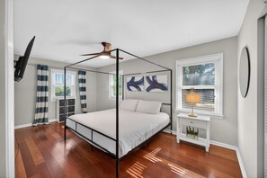 A bedroom with a four-poster bed, a ceiling fan, a TV mounted on the wall, and a window with blinds.