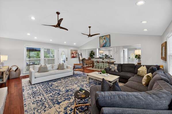 A spacious living room with a white sofa, a dark grey sectional, and a patterned rug. There are ceiling fans, a dining area, and a kitchen in the background.
