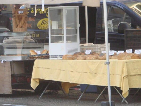 Un puesto de panadería con pan exhibido sobre una mesa, un refrigerador y un automóvil al fondo.