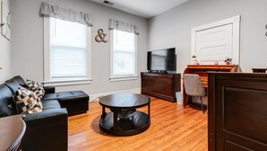 A living room with a black leather sofa, a round coffee table, a television on a wooden stand, and a wooden door. - Carytown Gem | Spacious Family-Friendly Apartment (Richmond)