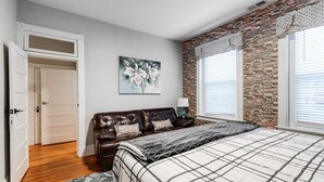 A bedroom with a bed, a brown leather sofa, a painting, and a stone wall. - Carytown Gem | Spacious Family-Friendly Apartment (Richmond)