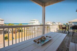 A covered outdoor area with a wooden table and chairs, overlooking a beach and several buildings.