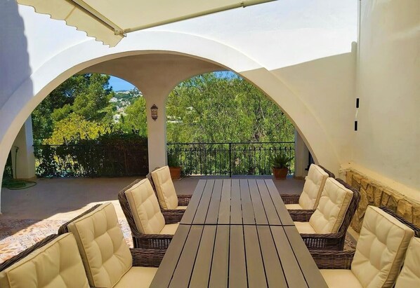 Une terrasse extérieure avec une table et des chaises, entourée d’arcs blancs, donnant sur un jardin avec des arbres et une clôture.
