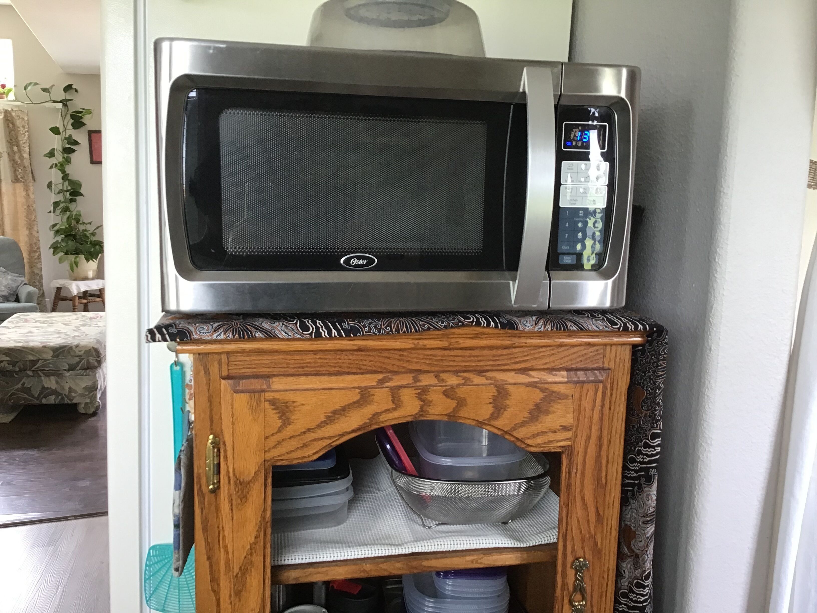 A microwave on a cabinet shelf, with a patterned tablecloth and a potted plant in the background.