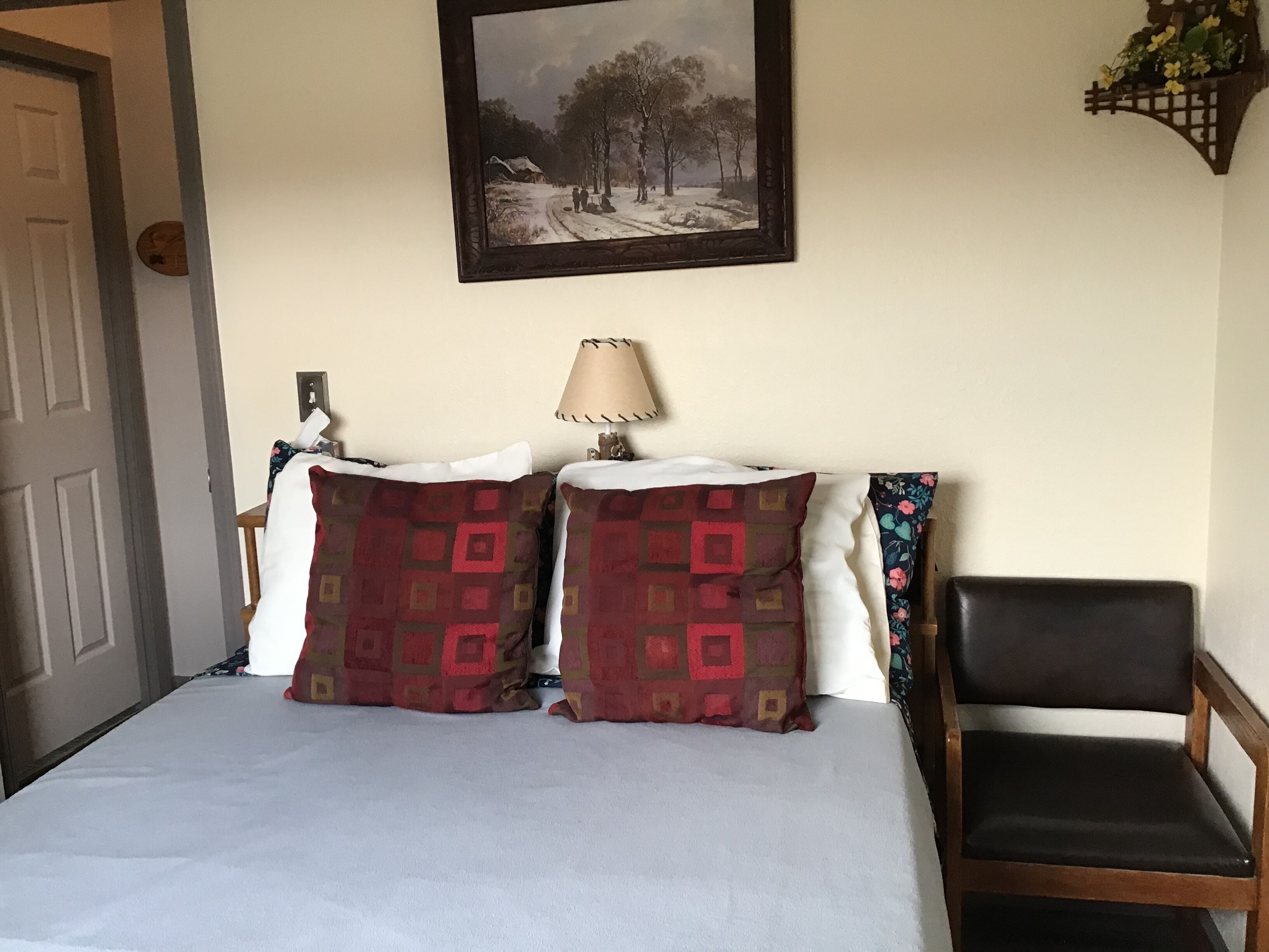 Een bed met gestreepte kussens, een nachtkastje met een lamp, een stoel en een schilderij van een besneeuwd landschap aan de muur.