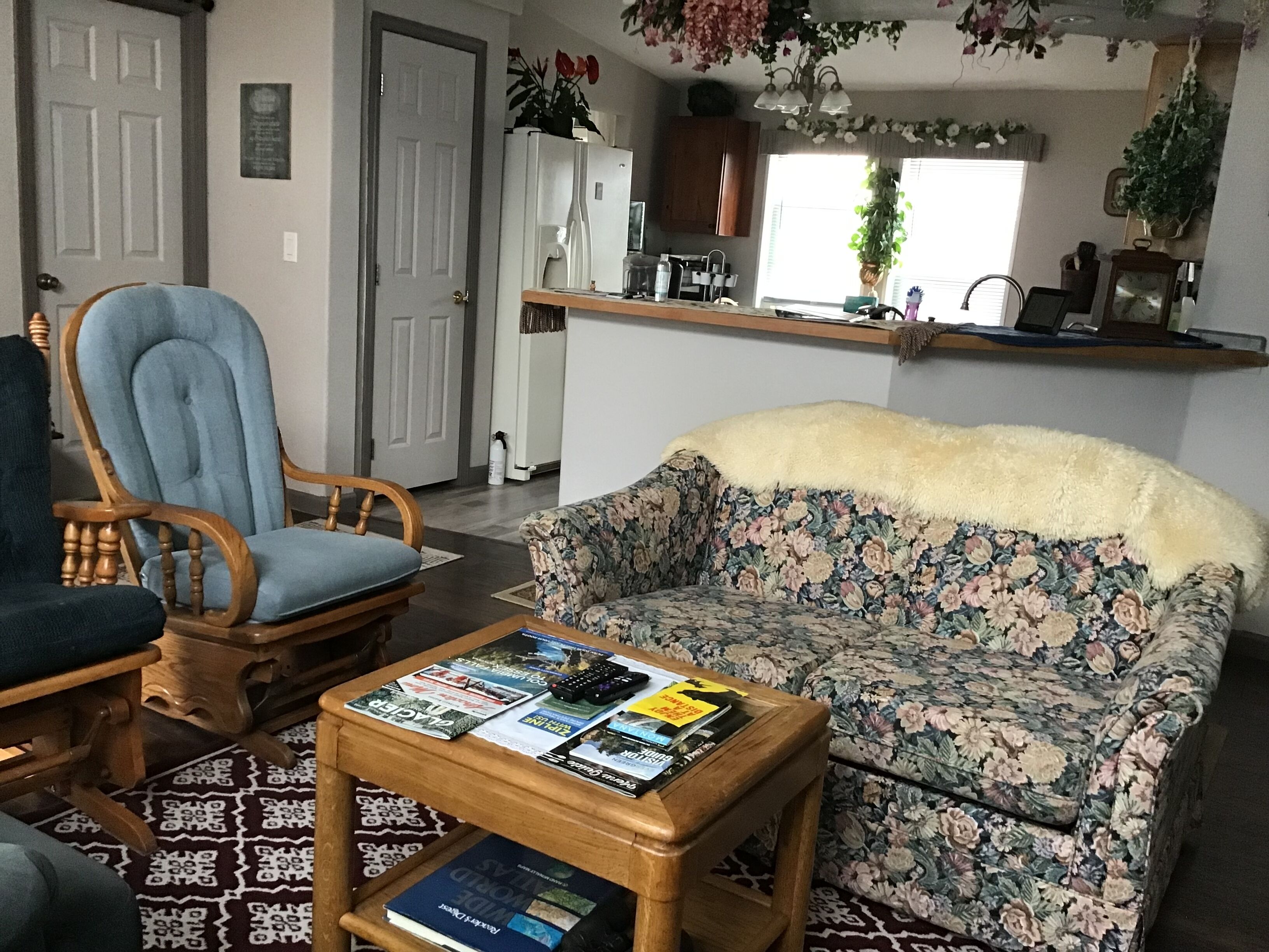 Un salon avec un canapé à motifs floraux, un fauteuil en bois, une table basse avec des magazines, et un coin cuisine à l’arrière-plan.