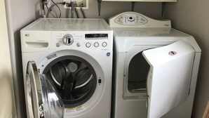 A laundry area with two washing machines, a control panel, and a shelf with a bottle and a box. - Bright and cheerful. With glorious sunrise/sunset views, close to Glacier park. (Columbia Falls)