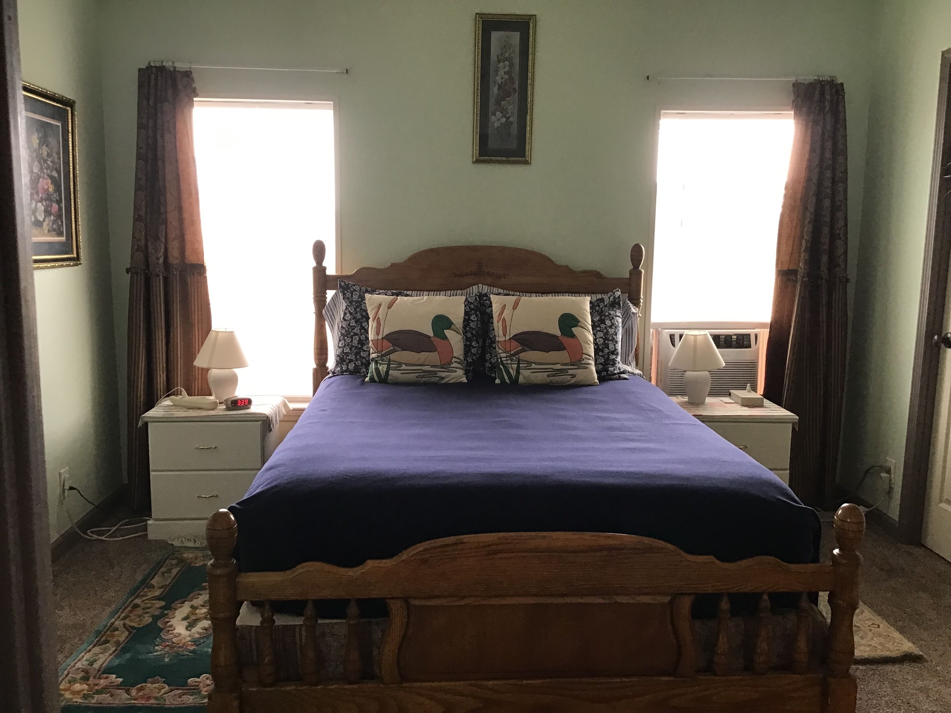 Un dormitorio con estructura de cama de madera, colcha morada, dos mesitas de noche con lámparas, un aparador y una alfombra estampada con motivos florales, además de una ventana con cortinas.