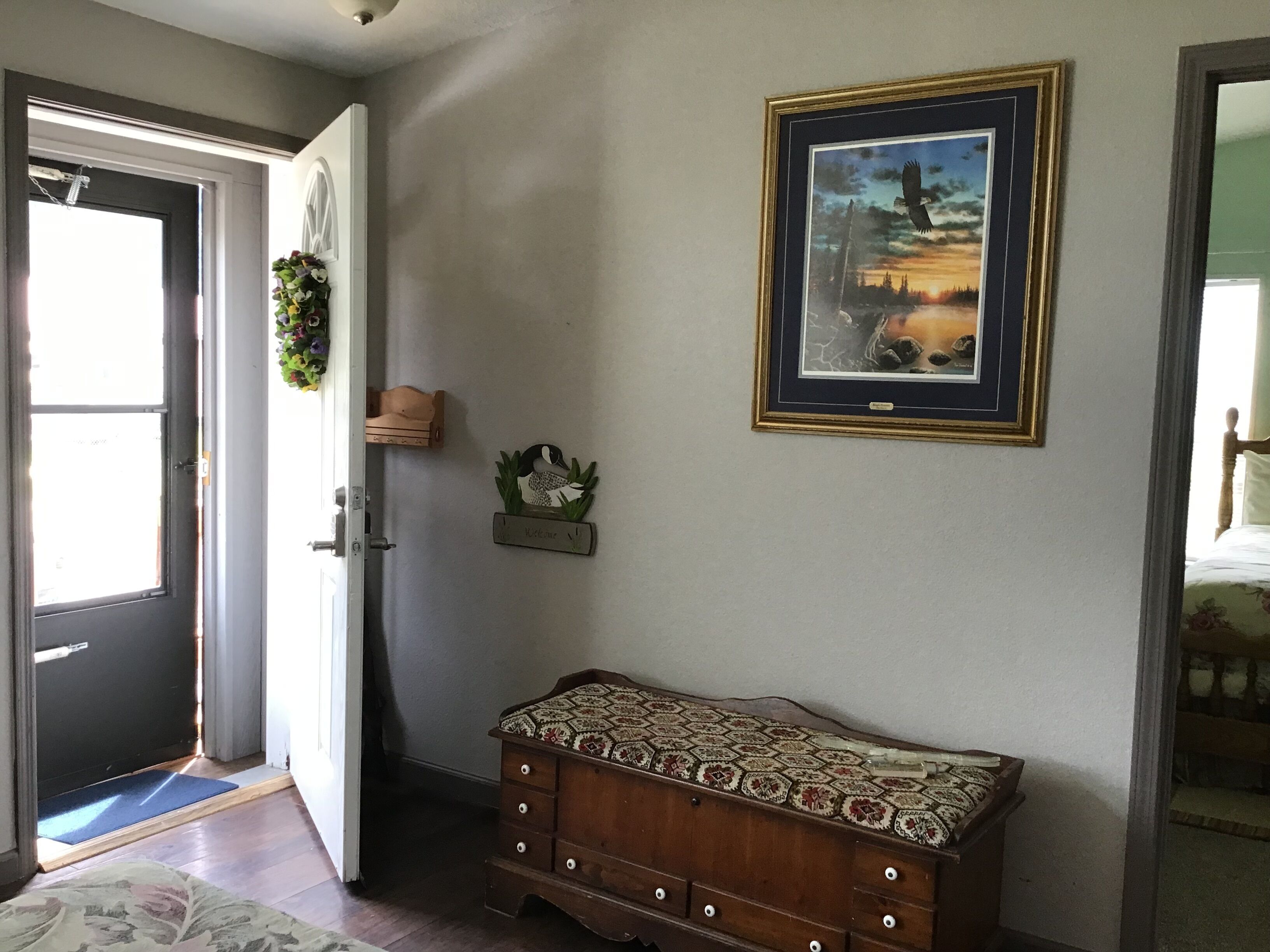 A room with a wooden bench, a framed picture of a landscape, a welcome sign, and a door with a mat.