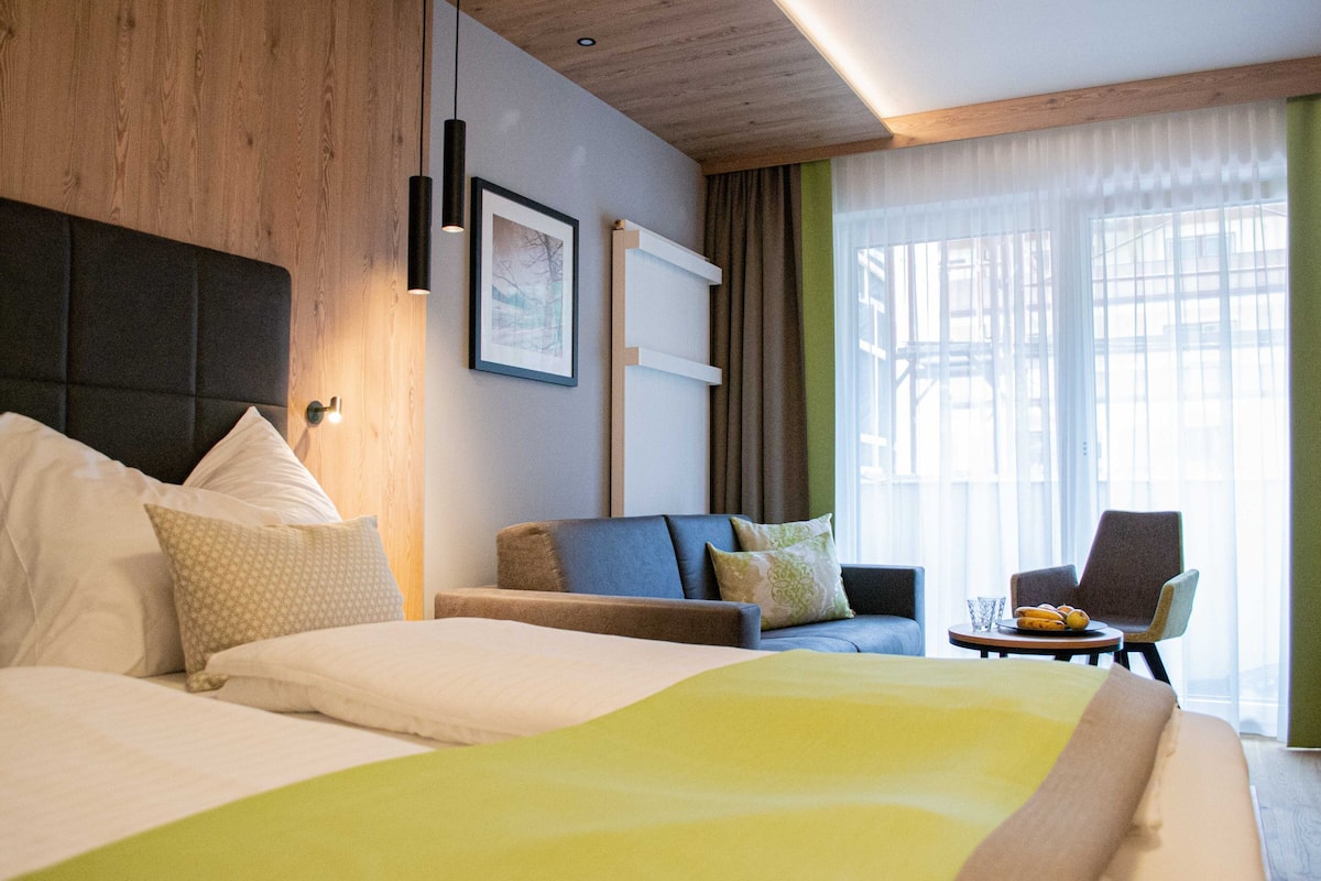 A hotel room with a large bed, a sofa, a chair, and a small table with a glass of water and a plate of fruit.