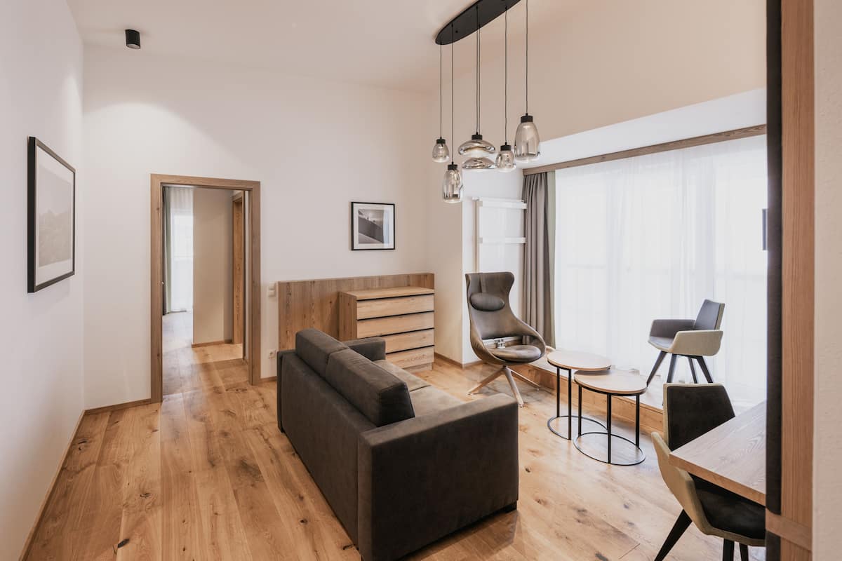 A modern living room with a sofa, a dining table with chairs, a large window with sheer curtains, and a wooden floor.
