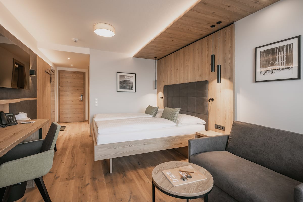 A modern hotel room with a wooden floor, a large bed, a sofa, a desk, and a small table.