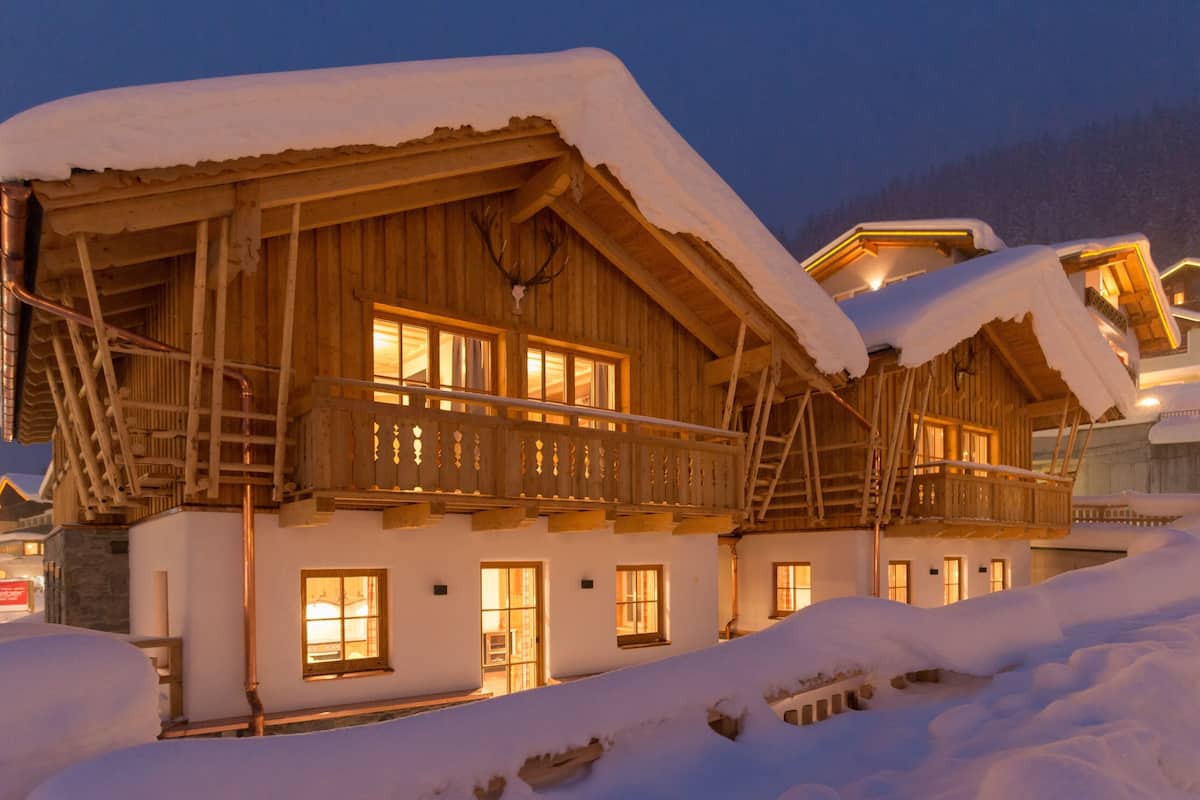 A wooden chalet with a balcony, snow-covered roof, and illuminated windows.