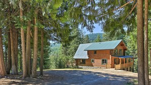 Une cabane en rondins surmontée d’un toit en métal, entourée de grands arbres et bordée d’une allée de gravier.