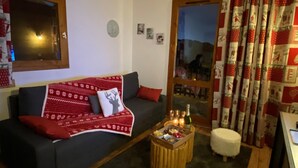 A cozy living room with a dark grey sofa, a red and white patterned blanket, a small wooden coffee table with a bottle and glasses, and a white fluffy ottoman. - Magnificent 6-person apartment 50 m from the slopes Household linen and towels included! (Valmeinier 1900)