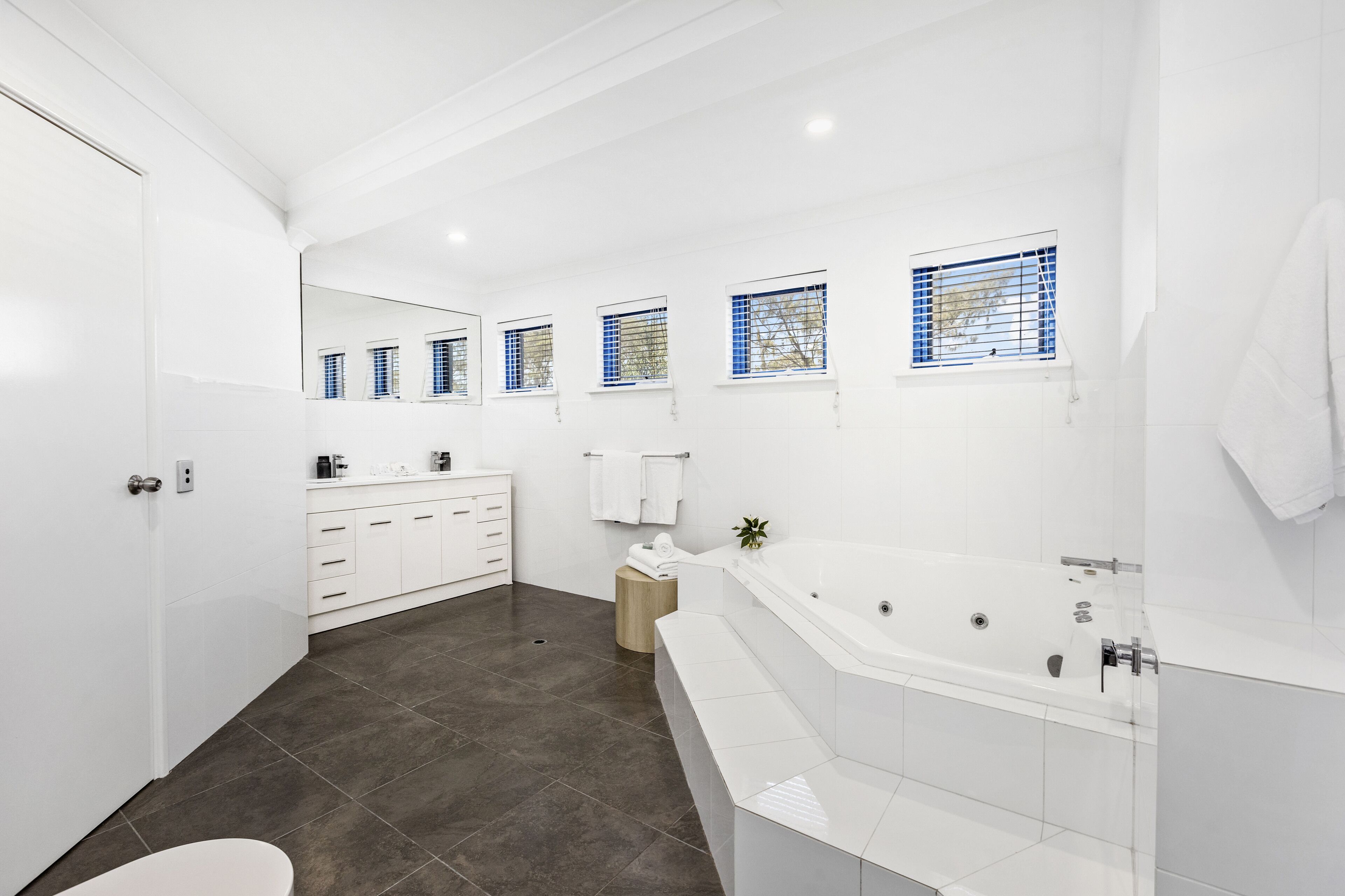 A modern bathroom with a large bathtub, a vanity with a mirror, and a window with blue blinds.