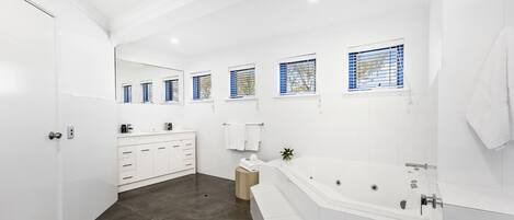 A modern bathroom with a large bathtub, a vanity with a mirror, and a window with blue blinds.