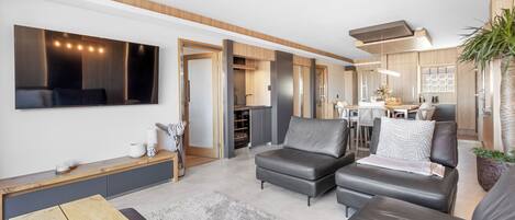 A modern living room with a flat-screen TV, leather sofas, a coffee table, and a dining area with a kitchen in the background.