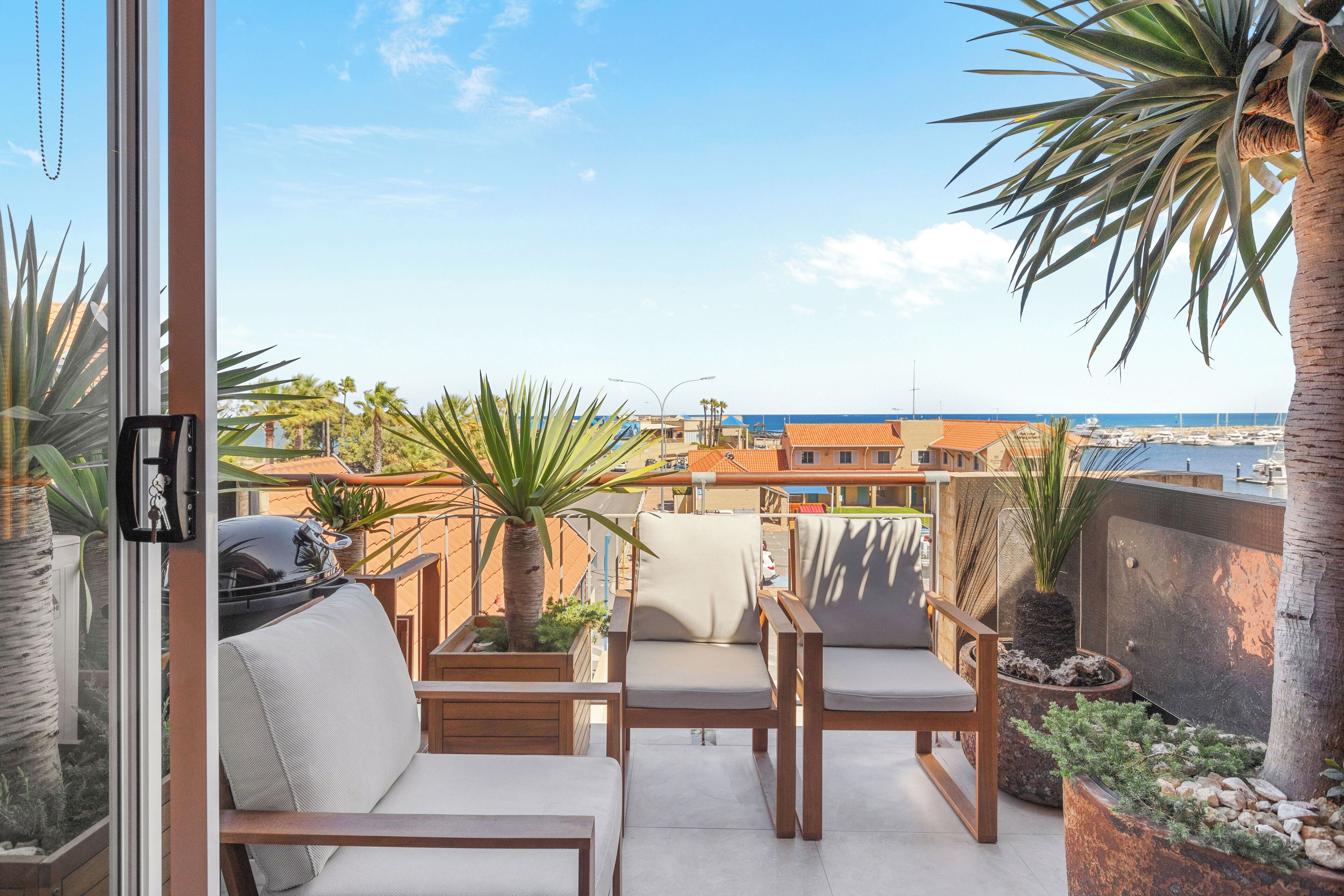 A balcony with a view of the sea, featuring a grill, seating area, and potted plants.