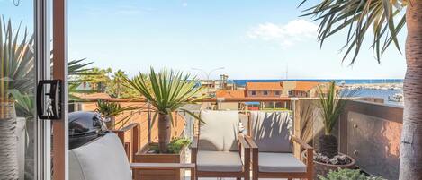 A balcony with a view of the sea, featuring a grill, seating area, and potted plants.