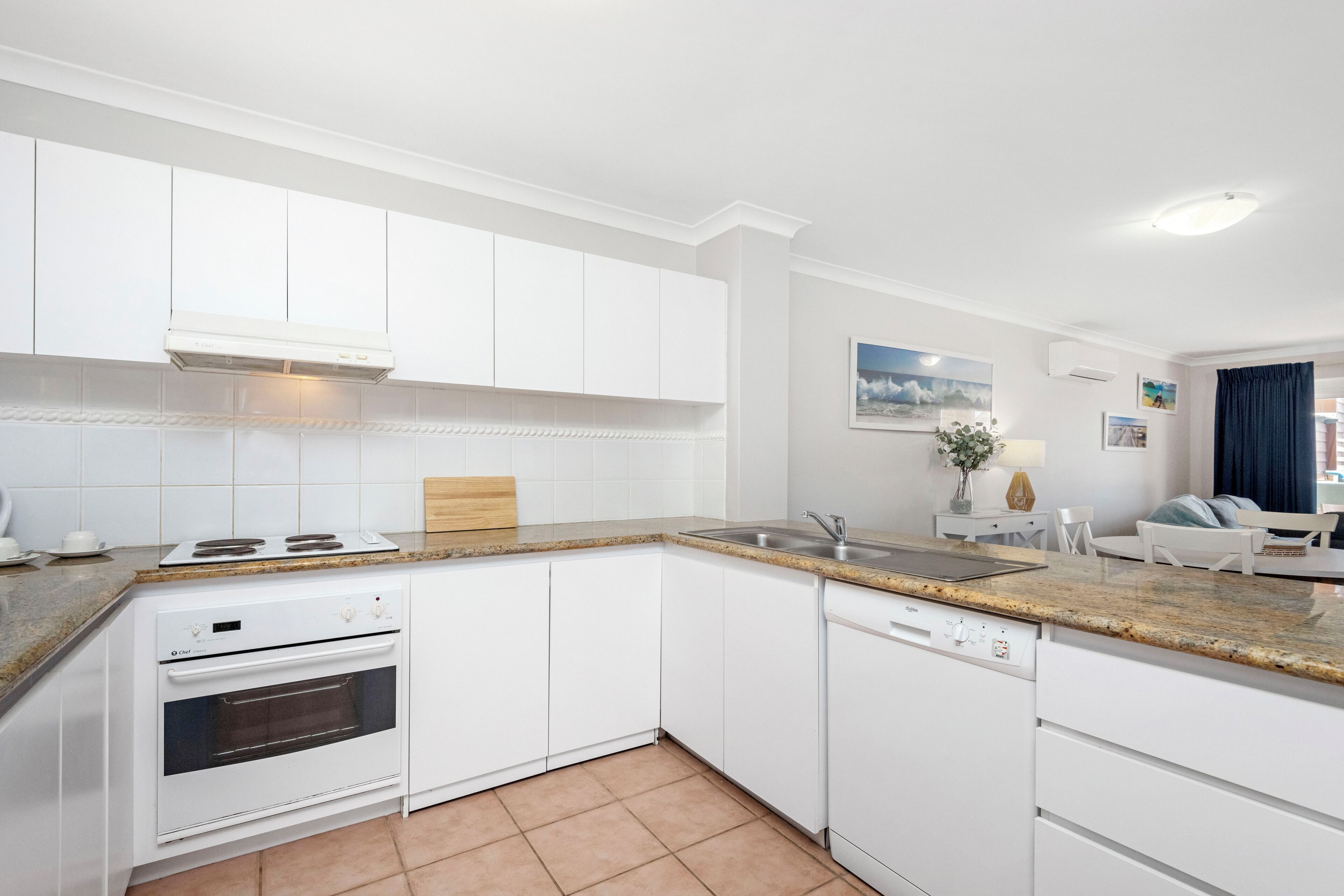 A modern kitchen with white cabinetry, a granite countertop, a built-in oven, and a dishwasher.