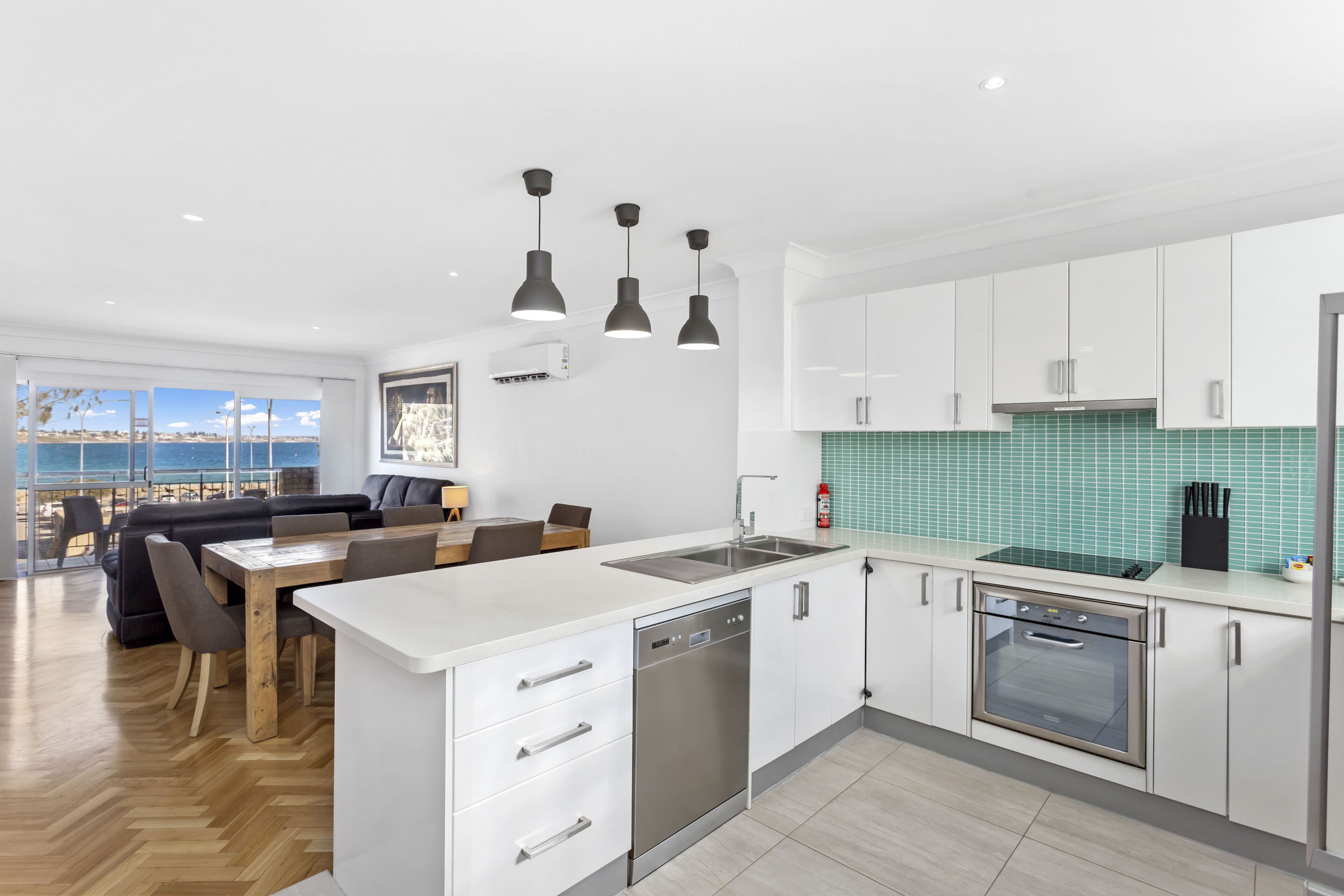 Een moderne keuken met witte kastjes, een roestvrijstalen vaatwasser en een oven. Er is een eethoek met een houten tafel en stoelen, en een groot raam met uitzicht op het strand.